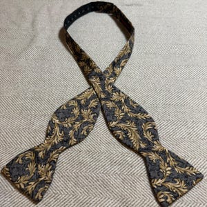 May include: A self-tie bow tie featuring a dark gray background with a repeating gold-colored acanthus leaf pattern. The fabric has a textured appearance, and the neck strap includes an adjustable metal slide mechanism for a secure fit.