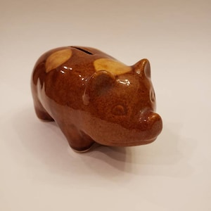 May include: A brown ceramic piggy bank with a slot on top for coins. The piggy bank has a light brown glaze with darker brown spots.