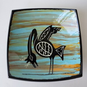 May include: A square ceramic dish with a black rim and a blue, yellow, and brown background. A black stylized bird with white details is painted on the dish.