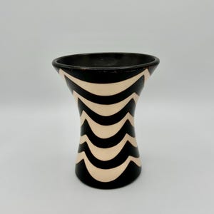 May include: A black and cream-colored vase with a wavy pattern. The vase has a flared top and a rounded base. The black and cream pattern creates a striking visual contrast, making it a decorative piece.
