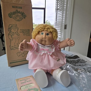 May include: A Cabbage Patch Kids doll with blonde yarn hair and green eyes, wearing a pink and white striped dress with a white collar and pink bow. The doll includes a cardboard box, a plastic bag, and a birth certificate and adoption papers.