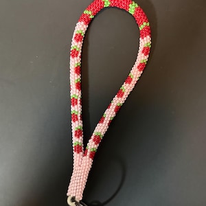 Op de afbeelding: Een polskoord gemaakt van kleine kraaltjes in een buisvormig patroon. Het ontwerp toont een herhalend motief van rode aardbeien met groene blaadjes op een lichtroze achtergrond. Een zilverkleurige metalen karabijnhaak en sleutelring zijn bevestigd.
