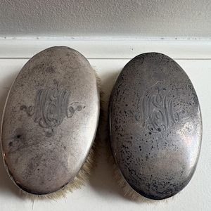 May include: Two oval-shaped brushes with tarnished silver-toned metal backs. Each brush features an intricate, engraved monogram in the centre of the metal surface and light-coloured bristles extending from the edges of the oval frames.