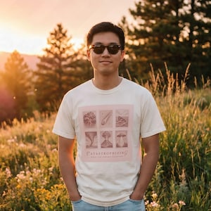 May include: A cream-coloured short-sleeve t-shirt featuring a square graphic print with six individual illustrations of natural disasters and the text "CATASTROPHIZING" and "WORST CASE SCENARIO COLLECTION" printed in a muted reddish-brown ink.