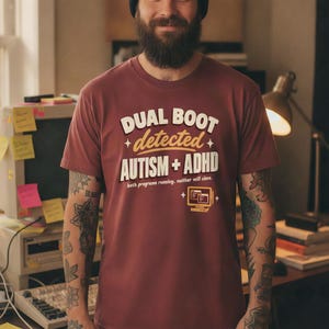Può includere: Una t-shirt a maniche corte color marrone con il testo bianco e giallo DUAL BOOT detected AUTISM + ADHD, il sottotesto both programs running. neither will close. e una piccola icona di computer gialla con due finestre.