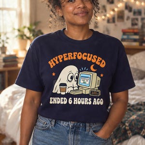 Op de afbeelding: Een marineblauw t-shirt met een afbeelding van een witte geest die met een koffiekopje achter een computer zit. De tekst luidt HYPERFOCUSED en ENDED 6 HOURS AGO, met een digitale klok die 3:00 am aangeeft en een scherm met HYPERFOCUS MODE: ON.