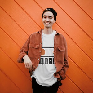 May include: A white t-shirt featuring a graphic print with the text LIQUID GLASS, worn under a rust-colored button-down shirt with two chest pockets, paired with a black beanie, showcasing a casual layered look against a bright orange background.