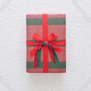 May include: A rectangular gift box wrapped in paper featuring alternating horizontal stripes of dark green and a textured reddish-brown wood grain pattern. The package is tied with a vibrant red ribbon that forms a centered bow.