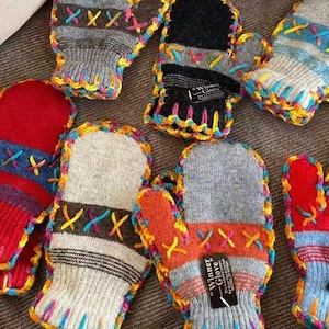 Könnte beinhalten: Eine Sammlung von Strickfäustlingen in verschiedenen Farben wie Rot, Grau, Schwarz und Orange. Jeder Fäustling ist mit dekorativen Kreuzstichen und einem bunten, gehäkelten Rand versehen. Einige Fäustlinge haben ein schwarzes Etikett mit der Aufschrift Winner Glove.