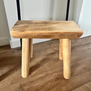 Può includere: Un piccolo sgabello rettangolare in legno con una finitura naturale color marrone chiaro. Lo sgabello è realizzato con una superficie superiore spessa e solida e quattro robuste gambe cilindriche che forniscono una base stabile al mobile.