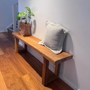 Op de afbeelding: Een lange houten bank met een natuurlijke houtnerfafwerking en vier stevige poten. Een lichtgrijs vierkant kussen ligt op de zitting en een groene bladplant in een bruine papieren zak staat aan de linkerkant op het oppervlak van de bank.