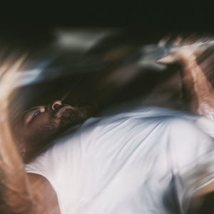 May include: A person with a beard wearing a plain white t-shirt, captured in a motion-blurred perspective while lying down. The image features a high-contrast, dark aesthetic with soft focus and light streaks across the frame, obscuring details.