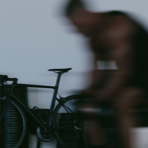 May include: A matte black road bicycle with thin tires and a streamlined frame, positioned in front of a stack of black circular weight plates. The bicycle features a standard saddle, drop handlebars, and a chainring, presented in a side profile.