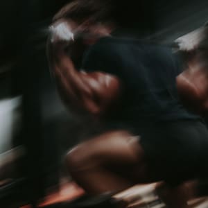 Könnte beinhalten: Eine unscharfe Bewegungsaufnahme eines Gewichthebers in schwarzem T-Shirt und schwarzer Shorts bei der Ausführung einer Kniebeuge mit Langhantel. Die Person ist mit der Hantel auf dem oberen Rücken positioniert und führt eine tiefe Kniebeuge aus.