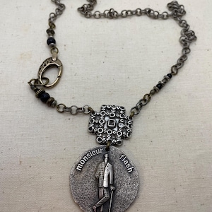 May include: Silver-toned necklace featuring a pendant with the words "monsieur flash" and an illustration of a man in a suit. The necklace has a chain with a decorative clasp and small black beads. The pendant is attached to a square, ornate silver piece.