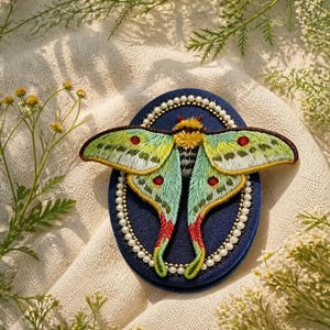 May include: An embroidered moth brooch featuring light green wings with red and white accents, a fuzzy yellow thorax, and a dark abdomen. The moth is mounted on a dark blue oval base bordered by a single row of small white beads.