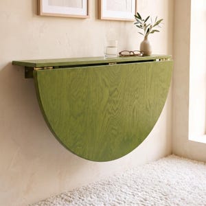 May include: A wall-mounted, semi-circular drop-leaf table crafted from olive green wood with a visible grain pattern. The table features gold-toned metal hinges and is shown in a folded position against a wall, with a glass and vase on top.