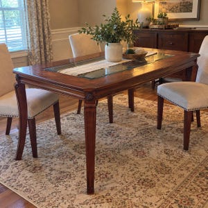May include: A rectangular wooden dining table with a central glass inlay, featuring carved corner details and straight legs. The table is surrounded by four upholstered chairs with light beige fabric and dark wooden legs.