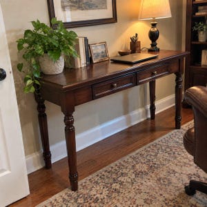 May include: A dark brown wooden writing desk featuring two drawers with round knobs and turned legs. The desk surface holds a potted green plant, several books, a small framed picture, a laptop, and a wooden pen holder with writing utensils.