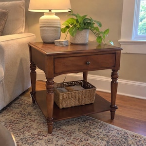 May include: A square wooden end table with a warm brown finish, featuring a single drawer with a small dark knob, four turned legs, and a lower shelf. A woven wicker basket sits on the bottom shelf, holding a book with a visible cover.