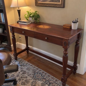 May include: A rectangular wooden writing desk with a dark brown finish, featuring two drawers with small round knobs, fluted legs, and an oval-shaped inlay design on the center of the tabletop surface.