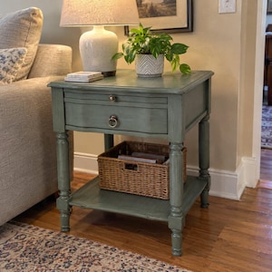 May include: A sage green wooden end table featuring a single drawer with a round metal ring pull and a lower shelf. A woven wicker basket sits on the bottom shelf. The table has four turned legs and a smooth, painted finish with subtle distressing.