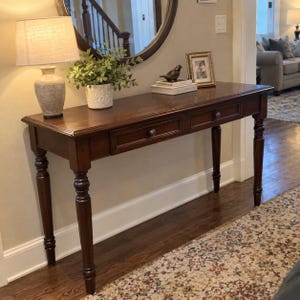 May include: A dark brown wooden console table featuring two drawers with small round knobs and four turned legs. The rectangular tabletop holds a beige lamp, a potted green plant, a small bird figurine, and a framed photograph.