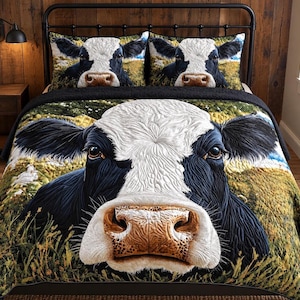 May include: A bedding set featuring a quilt and two pillow shams with a large, detailed print of a black and white cow face. The cow is set against a green pasture background. The quilt has a textured, quilted surface with a black border.