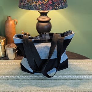May include: A tote bag featuring a color-blocked design of black and light blue denim fabric panels. The bag includes two black woven fabric handles and a black and white checkered interior lining visible at the top opening of the bag.