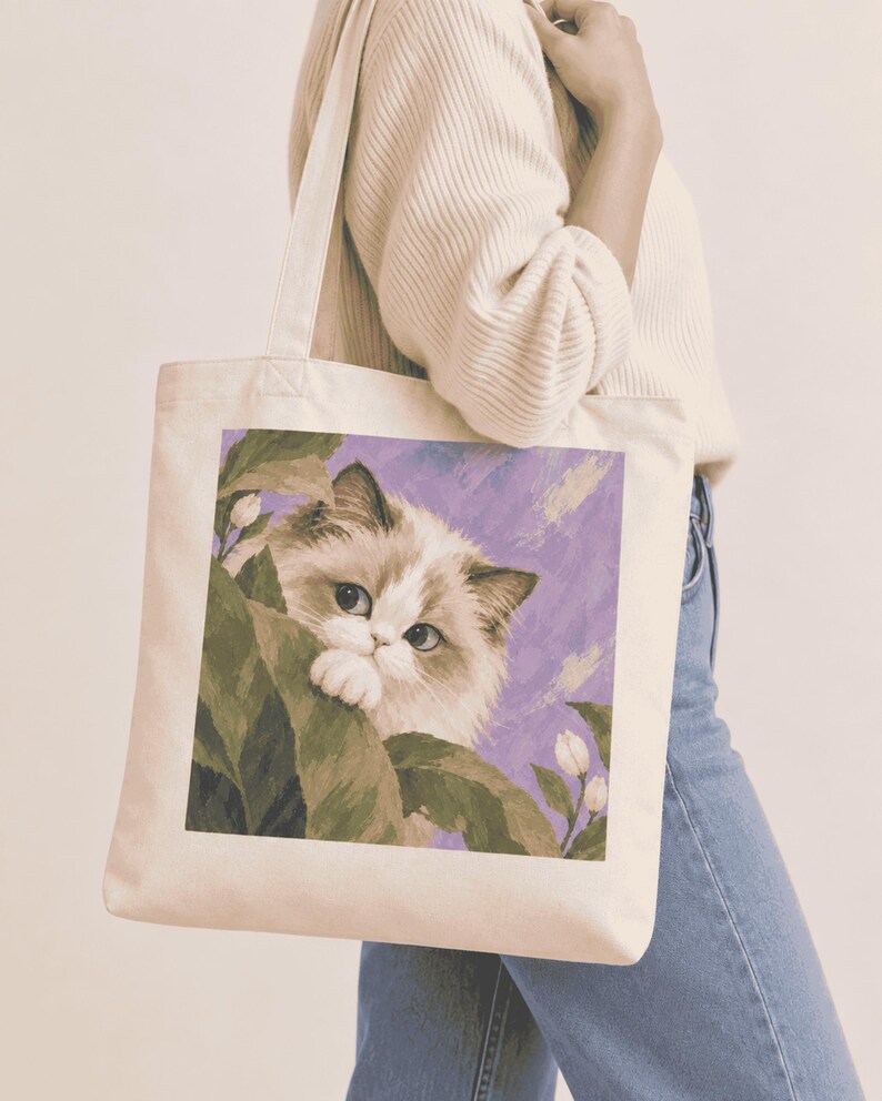 Canvas tote bag mockup featuring a long-haired cat among green leaves on a purple background.