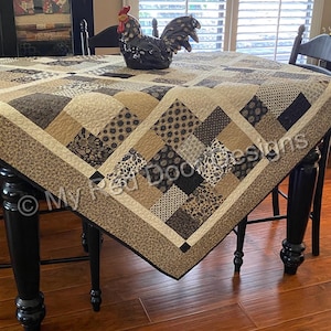 Op de afbeelding: Een patchwork quilt met een herhalend patroon van vierkanten in tinten beige, cr&egrave;me, bruin en zwart met bloemen- en geometrische prints. De quilt ligt over een tafel en bevat de tekst My Red Door Designs gedrukt op de stof.