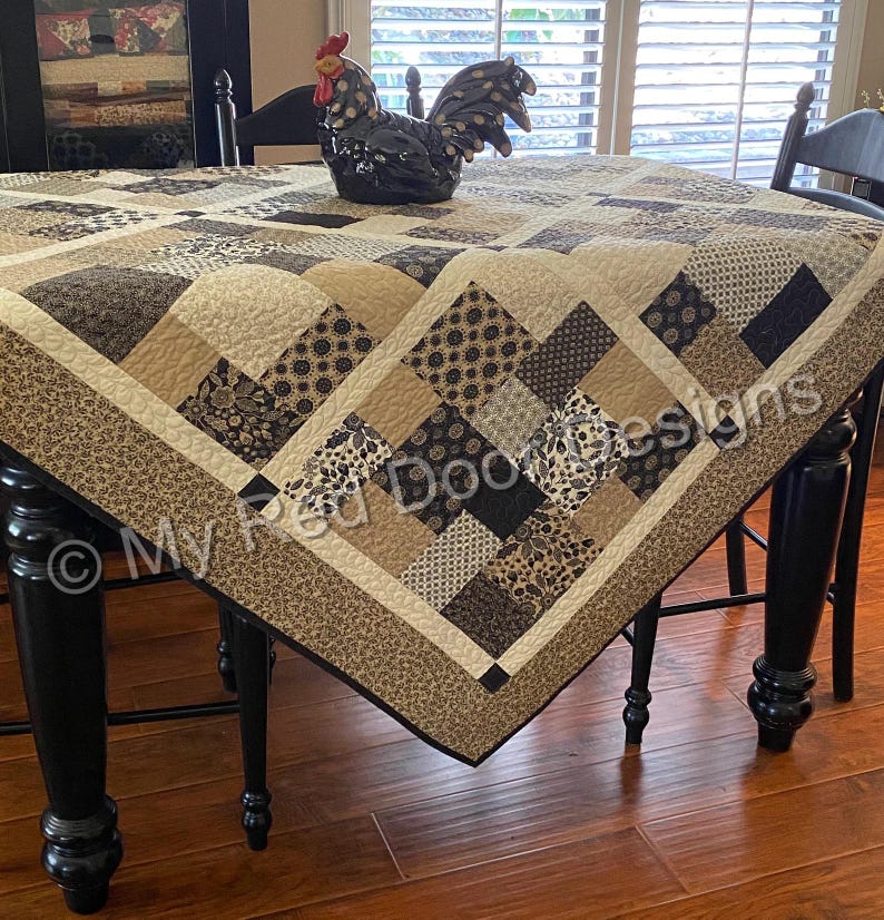 Op de afbeelding: Een patchwork quilt met een herhalend patroon van vierkanten in tinten beige, cr&egrave;me, bruin en zwart met bloemen- en geometrische prints. De quilt ligt over een tafel en bevat de tekst My Red Door Designs gedrukt op de stof.