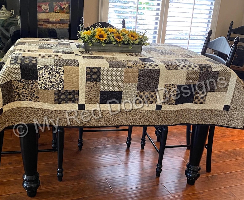 Op de afbeelding: Een rechthoekige quilt met een patchworkontwerp van diverse vierkante en rechthoekige stoffen blokken in tinten zwart, cr&egrave;me en beige. De quilt toont diverse patronen, waaronder bloemmotieven, geometrische vormen en paisley-prints met een rand.