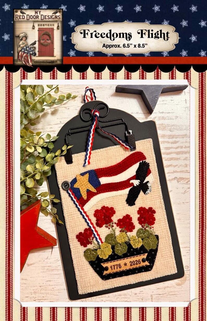 May include: A rectangular fabric wall hanging featuring a cream-colored background with a red, white, and blue flag, a black eagle, and red flowers in a black planter. The planter displays the text 1776 2026. It is held by a black metal clip.