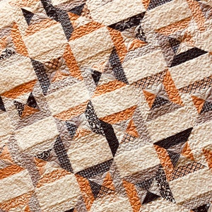 Puede incluir: Una colcha con un patr&oacute;n geom&eacute;trico repetitivo de bloques de tela en color crema, naranja y negro. Las secciones crema tienen una superficie acolchada texturizada y ondulada, mientras que las secciones oscuras incorporan varios patrones, incluyendo pata de gallo y floral.