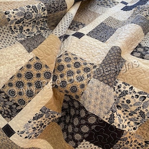 Op de afbeelding: Een patchwork quilt bestaande uit diverse vierkante stoffen blokken in tinten zwart, wit en beige. De quilt toont uiteenlopende patronen zoals bloemmotieven, geometrische vormen en bladeren, met zichtbare machinale stiknaden over het geheel.