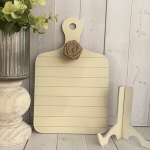 May include: A light beige wooden cutting board with a burlap flower accent. The board has a handle and is sitting on a white wooden surface.