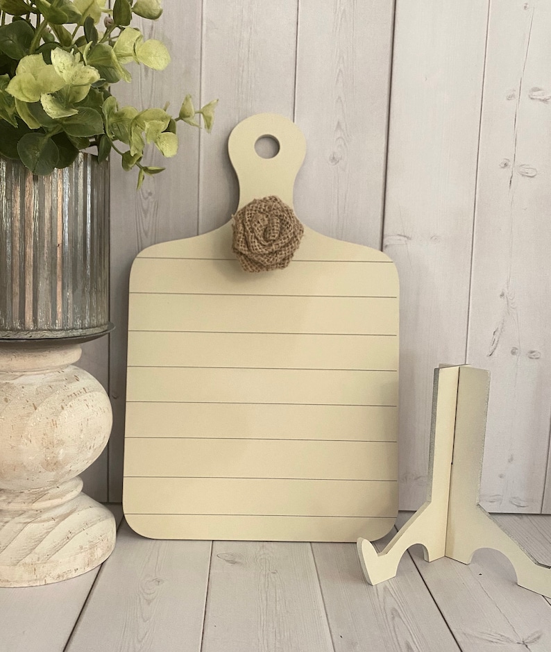 May include: A light beige wooden cutting board with a burlap flower accent. The board has a handle and is sitting on a white wooden surface.