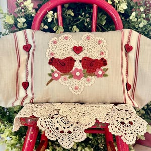 May include: A beige cushion with a heart-shaped design featuring two red birds and pink flowers. The cushion is on a red wooden chair with a crocheted doily.