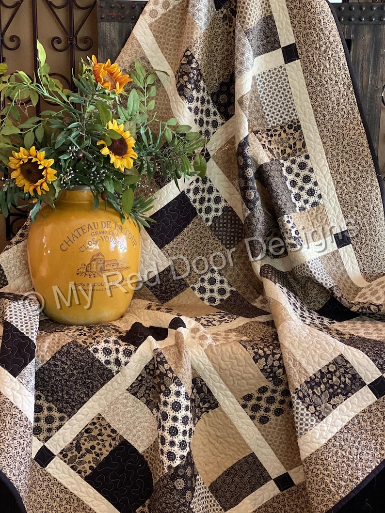 Op de afbeelding: Een patchwork quilt met diverse bruine, beige en cr&egrave;mekleurige stoffen vierkanten met bloem- en geometrische patronen, geaccentueerd door een gele keramische kan met zonnebloemen en de tekst My Red Door Designs op de voorzijde.