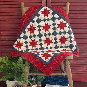 Könnte beinhalten: Eine patriotische Steppdecke mit einem rot-weiß-blauen Sternmuster. Die Steppdecke zeigt rote Sterne auf einem weiß-blauen karierten Hintergrund, umrahmt von einem roten Rand. Sie ist über einen Holzstuhl drapiert.