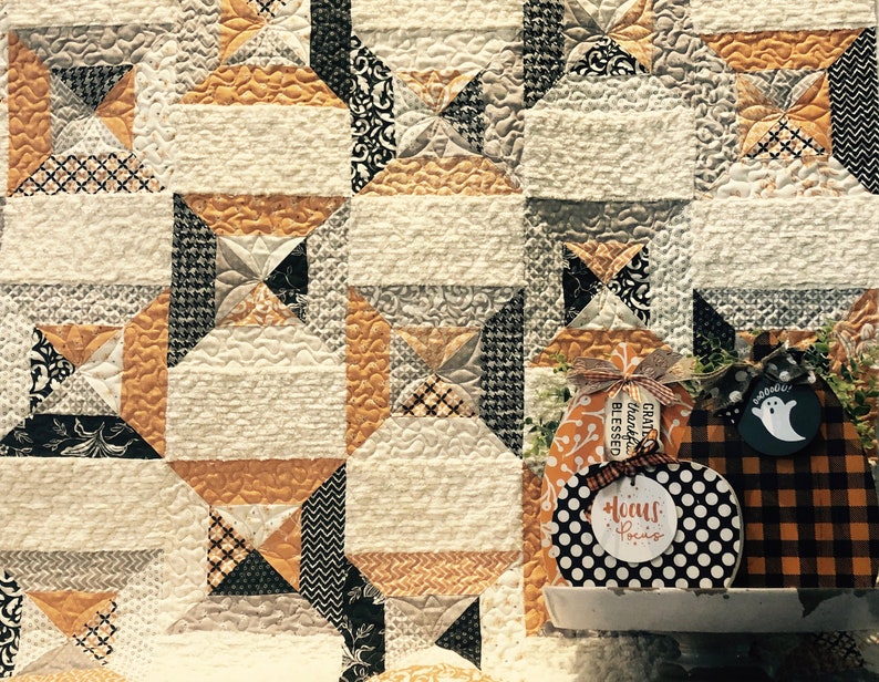 Puede incluir: Un fondo de tela acolchada con un patr&oacute;n geom&eacute;trico en crema, naranja y negro. En primer plano, se muestran dos decoraciones de madera con forma de calabaza. Una presenta lunares blancos y negros con el texto "hocus pocus".