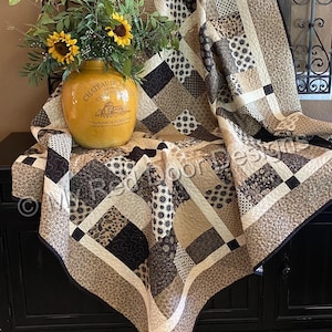 Op de afbeelding: Een patchwork quilt met een verscheidenheid aan bruine, beige en cr&egrave;mekleurige stoffen vierkanten met diverse bloem-, geometrische en paisley-patronen. De quilt is over een oppervlak gedrapeerd en toont ingewikkelde stikselpatronen.