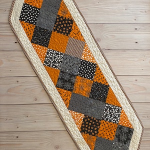 May include: A Halloween-themed table runner with a patchwork design featuring orange, black, and grey squares with various Halloween-themed patterns. The runner has a white border with a brown trim.