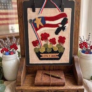 May include: A wooden display stand holding a rectangular fabric wall hanging featuring a red, white, and blue flag, a bald eagle, and red flowers. The fabric includes the text 1776 * 2026. A small leather-bound book and a metal pen sit on the base.