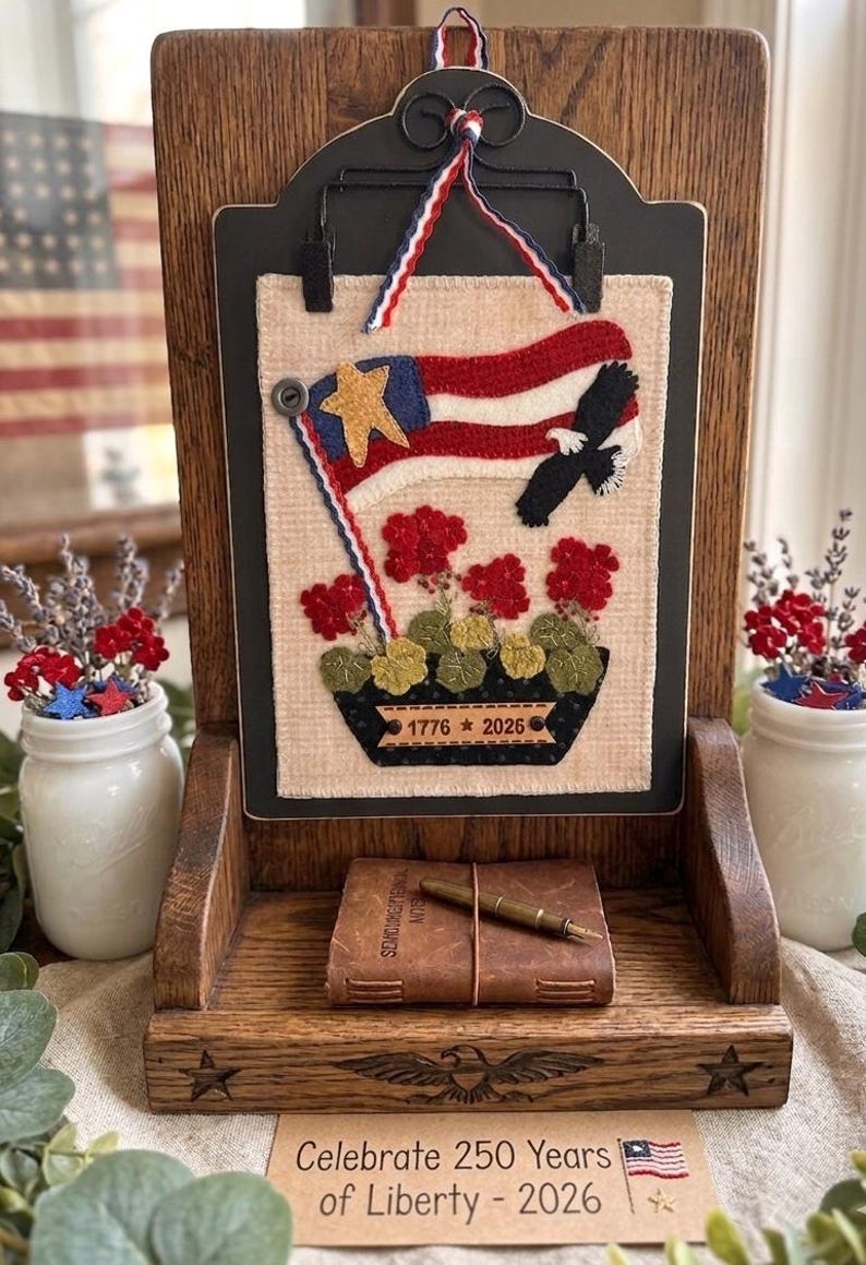 May include: A wooden display stand holding a rectangular fabric wall hanging featuring a red, white, and blue flag, a bald eagle, and red flowers. The fabric includes the text 1776 * 2026. A small leather-bound book and a metal pen sit on the base.