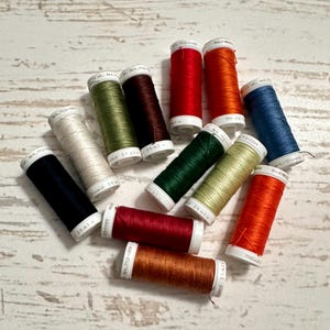 Puede incluir: Una colección de coloridas bobinas de hilo en varios tonos, incluyendo rojo, naranja, verde, azul y marrón. Las bobinas están dispuestas sobre una superficie de madera clara, mostrando los vibrantes colores del hilo.