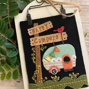 May include: A felt wall hanging with a black background featuring a vintage camper, a sign that reads "Happy Campers", and a red bird. The camper is light blue with white and orange accents. The sign is brown with white lettering. The background is black with green grass and yellow flowers.