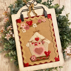 May include: A felt wall hanging with a birdhouse design. The birdhouse is white with a pink heart and red buttons. There are two red birds and pink flowers on a branch above the birdhouse. The background is burlap with a brown and red scalloped border.