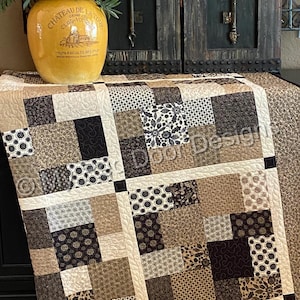Op de afbeelding: Een patchwork quilt met een raster van vierkante stoffen blokken in tinten zwart, bruin, beige en cr&egrave;me. De quilt toont diverse patronen waaronder bloemmotieven, geometrische vormen en krulontwerpen, gescheiden door cr&egrave;mekleurige stroken.